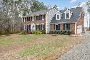 A two-story brick house with dark shutters, surrounded by a lightly wooded area. The driveway is gravel, and the lawn is slightly brown. The sky is clear, and trees border the property.