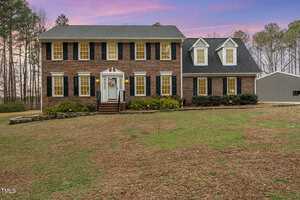 A two-story brick house on 2000 Hayes Road features a large front lawn at sunset. With black shutters, a white door, and dormer windows, it's enveloped by trees. The property in Creedmoor also includes a detached gray garage visible to the right.