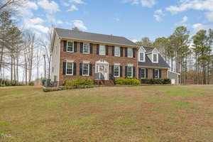 A two-story brick house with dark shutters and a front porch situated on a large grassy plot. Trees surround the property, and the sky is partly cloudy. There is a semi-circular driveway leading to the front of the house.