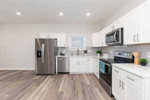 Modern kitchen with white cabinets, stainless steel appliances including a refrigerator, oven, and dishwasher. Light wood flooring complements the clean design. Small plants decorate the countertop for a touch of greenery.