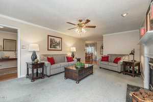 A cozy living room with two gray sofas adorned with red pillows. There's a central wooden coffee table, lamps, and a ceiling fan. A painting hangs on the wall, and a fireplace is in the corner. The room is warmly lit and inviting.