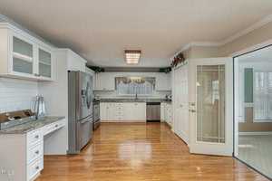 A spacious kitchen with white cabinets, granite countertops, and stainless steel appliances, including a fridge and dishwasher. There's a tile backsplash, hardwood floors, and a decorative border above the cabinets. Glass doors open to an adjacent room.