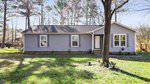Located at 903 W C Street in Butner, a single-story gray house with a dark roof stands in a grassy yard. A tree graces the foreground, and a wooden fence encloses part of the backyard. The house features three visible windows and a central front door, with trees lining the background.