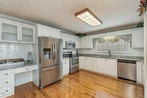 A kitchen with white cabinets, stainless steel appliances, and a granite countertop. It features a large window with a floral valance. The hardwood floor adds warmth to the space, and a rectangular ceiling light illuminates the room.