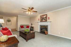 A cozy living room with a ceiling fan and fireplace. The room features a gray sofa adorned with red and gray pillows, a wooden coffee table with decorative items, and a framed painting above the mantel. The carpet is a soft green color.