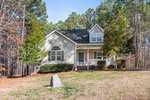A light-colored house with a small porch and an American flag sits serenely at 4078 Brooksdale Drive in Franklinton. The front yard features grass and some shrubs, with a stone marker visible. Tall trees envelop the home, creating a peaceful wooded setting.