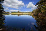 Nestled near 00 Uzzle Road in Rougemont, a serene lake reflects a bright blue sky with fluffy clouds, surrounded by lush green trees and a grassy landscape. This peaceful setting suggests a quiet, tranquil day immersed in nature's beauty.