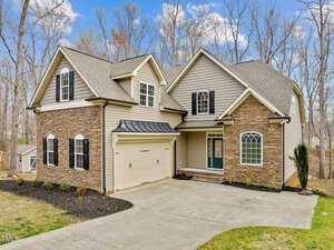 A two-story suburban house with beige siding and stone accents. It has a double garage, black shutters, and a front porch. The yard is partially landscaped, and the background features leafless trees and a clear blue sky.