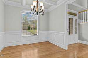 Elegant empty room with hardwood floors, light green walls, and white wainscoting. A large window with blinds overlooks a garden. A black chandelier hangs from a coffered ceiling, and a white door with glass panels is on the right.