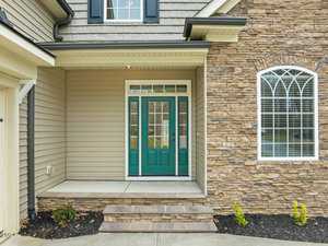 Front porch of a house with stone and siding exterior, featuring a green door with glass panels and two sidelights. An arched window is on the right above stonework, and the entrance is accessed via a small step with a concrete path.