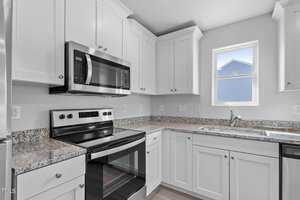 A modern kitchen with white cabinets and granite countertops. It features a stainless steel electric stove and microwave, a dishwasher, and a sink beneath a window. The space is well-lit and spacious.