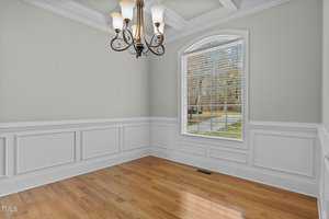 A bright, empty room with light green walls and white wainscoting. There's a large arched window with blinds, a hardwood floor, and a ceiling-mounted chandelier with multiple lights.