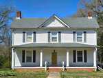 Located at 218 US Highway 15 in Stovall, this two-story white house with gray shutters and a gabled roof features a charming covered front porch supported by white columns. A central door and two chimneys complete the picturesque setting against a backdrop of trees and a clear blue sky.
