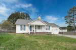 A white single-story house with a gray roof and front porch at 2753 Joyce Court, set against a blue sky. This Creedmoor residence features a brick foundation, well-maintained lawn, and a driveway on the right. Trees and a wooden fence add charm to the background.