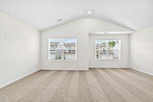 A spacious, empty room with light-colored walls and carpeted floor. Two windows provide natural light, overlooking neighboring houses. The ceiling is vaulted, adding to the airy feel of the space.