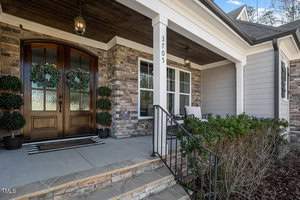 A welcoming house entrance with double wooden doors adorned with wreaths. The porch features stone accents, a black railing, neatly trimmed shrubs, and an outdoor lantern, creating a cozy and inviting atmosphere. House number "3705" is displayed.
