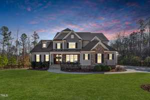Suburban two-story house at 3705 Dillard Lane with a gabled roof and stone facade, warmly lit windows against a twilight sky. Nestled in serene Wake Forest, it is surrounded by manicured lawns and trees, creating an inviting atmosphere.