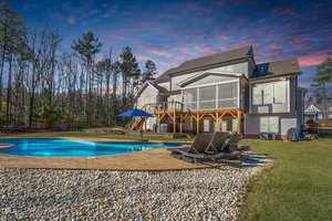 A two-story house with a large deck overlooks a backyard featuring a round swimming pool surrounded by a stone patio. Three lounge chairs are positioned by the pool. Trees and a neighboring house are in the background, under a colorful, cloudy sky.
