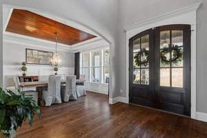 A modern dining room with light gray walls and a wooden floor. There's a long table with covered chairs and a spherical chandelier. The room opens into a foyer with double dark wooden doors, and a large plant is in the corner.