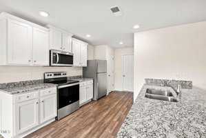 A modern kitchen with white cabinets, granite countertops, stainless steel appliances including a refrigerator, oven, and microwave. The room has hardwood floors and a double sink on the island countertop.