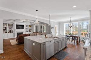 A modern kitchen and living room with a large marble island, pendant lights, and wooden flooring. The kitchen opens to a dining area with large windows, providing natural light. A cozy living area has a wall-mounted TV and built-in shelves.