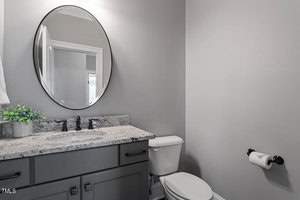 A modern bathroom with gray walls features a round mirror above a gray granite countertop. It has a sink with black fixtures, a white toilet, and a toilet paper holder. A small potted plant decorates the countertop.