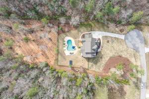 Aerial view of a large house with a gray roof surrounded by trees. The property features a swimming pool in a fenced backyard, a driveway, and a circular cul-de-sac on the right. The landscape is a mix of brown and green.
