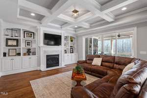 Spacious living room with a brown leather sectional, a fireplace, built-in shelving, and a TV. Large windows and glass doors lead to a porch. The room features a coffered ceiling and hardwood floors. A coffee table with a plant is centered on a rug.
