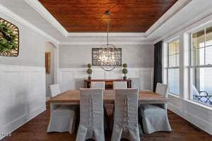 A dining room with a wooden table and six gray slipcovered chairs. A modern chandelier hangs from a wooden ceiling. There are large windows with white trim, a sideboard, greenery, and framed artwork on the walls.