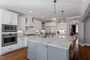 A modern kitchen featuring white cabinetry, stainless steel appliances, a large gray island with a granite countertop, and pendant lighting. Wooden bar stools line the island, and a vase of yellow flowers adds a touch of color.