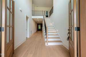 A spacious entrance hallway with light wooden flooring, double wooden doors, and a staircase with white risers and wooden treads leading to an upper floor. The walls are white, and a railing overlooks the entrance from above.