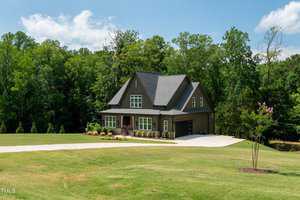 A two-story dark gray house with a steep roof, large windows, and a spacious driveway is surrounded by lush green trees and a manicured lawn under a partly cloudy sky.
