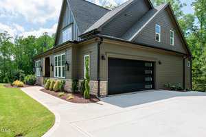 A modern two-story house with gray siding, large windows, and a black garage door. The driveway is wide and clean, and the garden is landscaped with small bushes. Trees and a clear sky are in the background.