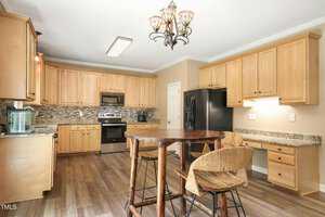 A spacious kitchen with light wood cabinets, granite countertops, and stainless steel appliances. The room features a wooden table with wicker chairs and a decorative ceiling light. The floor is made of dark wood, adding contrast to the space.