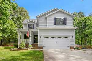 Nestled at 3715 Sapphire Court in Franklinton, this two-story gray house with white trim and a gabled roof exudes charm. Its inviting front porch, adorned with an American flag, complements a two-car garage on the right, all amidst lush green trees and a neatly trimmed yard.