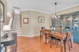 Dining room with a wooden table and chairs, a black side table with a lamp, and a glass-front cabinet. The walls are adorned with floral artwork. Stairs with wooden steps and white risers are visible in the background. A chandelier hangs above the table.