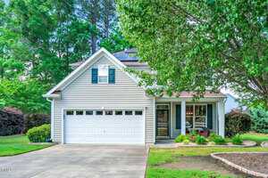 A suburban house with light gray siding and a two-car garage, surrounded by lush green trees and manicured grass. A concrete driveway leads up to the garage, and there's a small front porch with a bench.