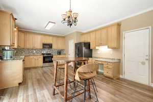 A spacious kitchen with light wood cabinets, granite countertops, and stainless steel appliances. A round wooden table with three matching chairs is centered on wood flooring. A decorative chandelier hangs above the table, and a door is visible in the back.