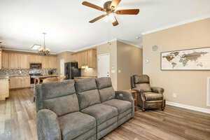 A living room with a gray sofa, recliner, ceiling fan, and a world map on the wall. The room leads into a kitchen with wooden cabinets, a black refrigerator, and a chandelier above the dining table. The floor is wood laminate.