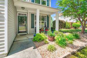 A charming white house with a covered front porch, featuring a black door and green shutters. The porch has potted plants and red chairs. The front yard includes a landscaped garden with rocks, shrubs, and a small tree.