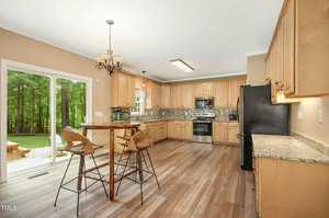 A spacious kitchen with wooden cabinets, granite countertops, and stainless steel appliances. A round high table with two wooden chairs sits in the center. Glass doors open to a lush green backyard.