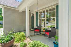 Front porch of a house with two black chairs featuring red cushions, a small table with yellow flowers, potted plants, white columns, and a large window. The porch overlooks a driveway and street lined with greenery. .