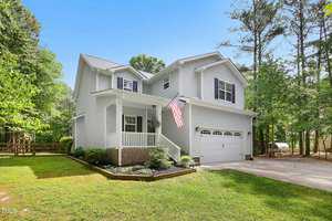 A two-story white house with a large front porch, an American flag, and a two-car garage. Surrounded by trees, the house sits on a well-maintained lawn with a paved driveway.