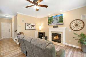 A cozy living room with a beige color scheme features a gray couch, a ceiling fan, and a fireplace beneath a wall-mounted TV. A map and wagon wheel decor adorn the walls. Natural light streams through a window, illuminating a potted plant.