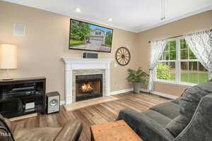 A cozy living room with a fireplace and mounted TV above it displaying a house exterior. There's a gray sofa, armchair, small wooden table, and a large window with patterned curtains. A plant sits in the corner, and the floor is wooden.