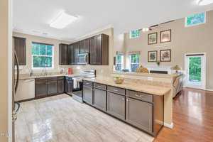 Modern kitchen with dark wood cabinets and granite countertops. Stainless steel appliances and a sleek design. Open layout leading to a spacious living area with large windows, artwork on the walls, and hardwood floors. Bright and inviting space.