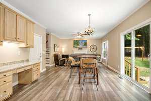 A cozy open living area featuring a kitchen with wooden cabinets and granite countertops. A small desk is built into the kitchen area. The dining table and chairs are situated near a glass sliding door, leading to a deck with a view of greenery outside.