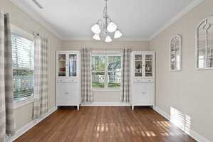 Bright dining room with wooden floor, two white cabinets with glass doors, and a window with patterned curtains. A chandelier hangs from the ceiling, and two decorative mirrors are on the right wall.