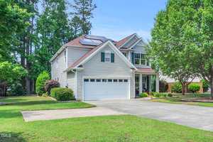 Located at 2104 Queensway Court in Creedmoor, this two-story suburban house features solar panels on the roof, a double garage, and a tidy front lawn. With white siding and trees and shrubs surrounding it, the home sits under a clear blue sky.