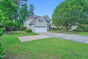 A two-story house with solar panels on the roof, a double garage, and a neatly manicured lawn. Trees and shrubs surround the property, and a driveway leads up to the garage. The sky is clear and blue.